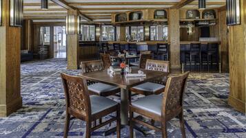 A dining area with a large wooden table surrounded by chairs, a patterned carpet, and a bar area with shelves and glassware.