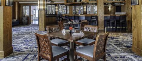 Un comedor con una gran mesa de madera rodeada de sillas, una alfombra estampada y una zona de bar con estantes y cristalería.