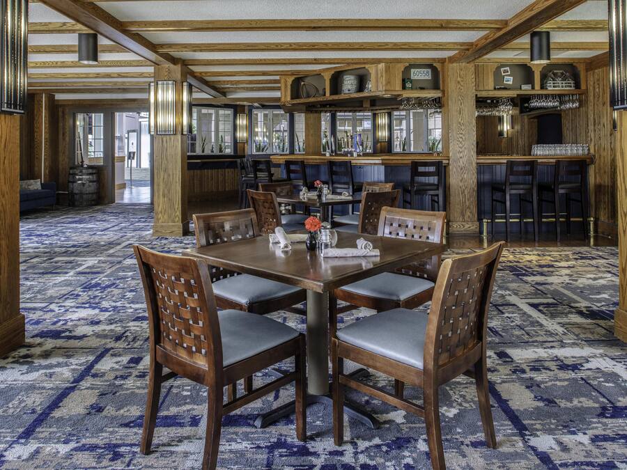 A dining area with a large wooden table surrounded by chairs, a patterned carpet, and a bar area with shelves and glassware.
