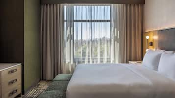 A hotel room with a large bed, a nightstand, a lamp, and a view of the outdoors through the window.