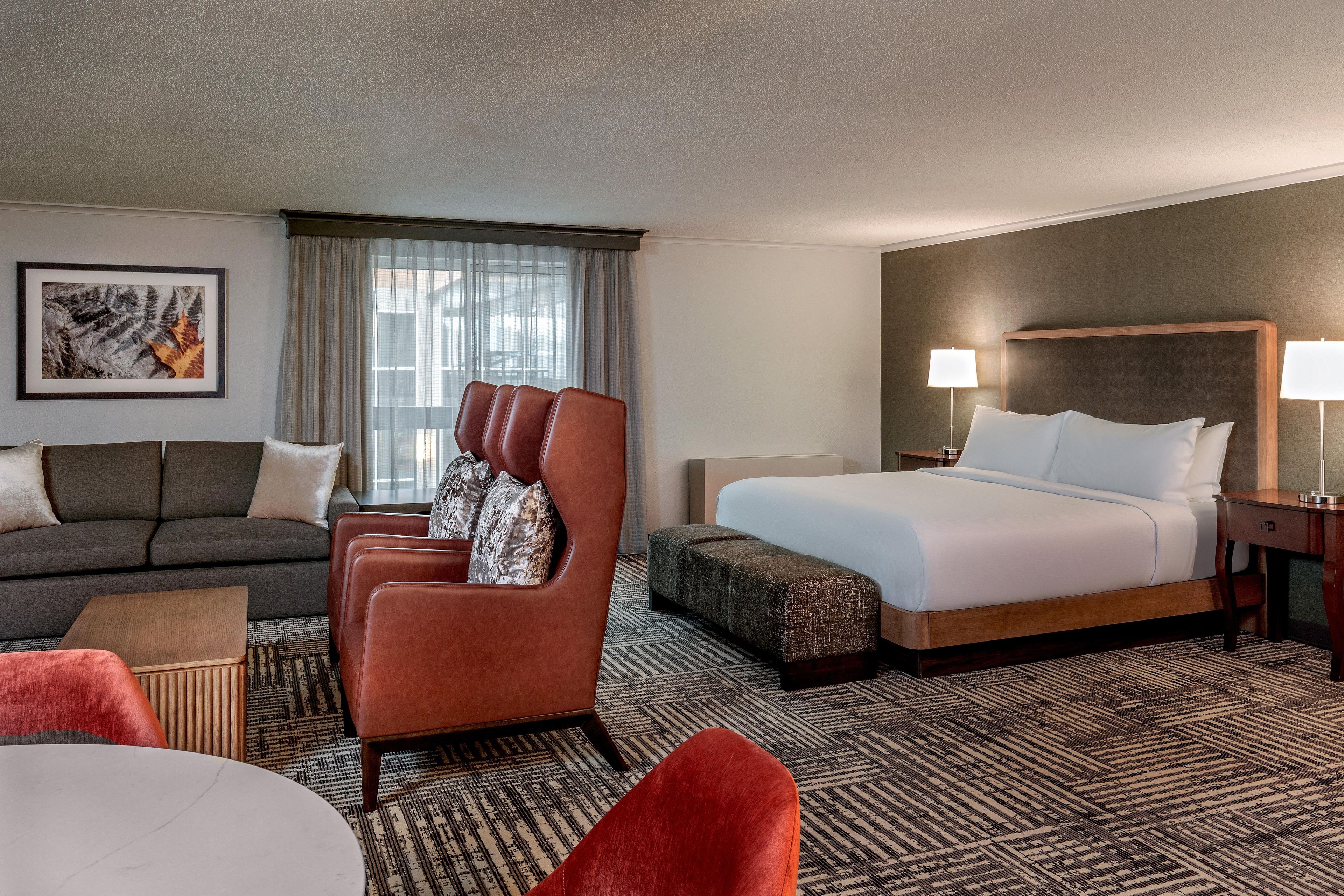 A hotel room with a sofa, a red armchair, a bed, a nightstand, and a wall-mounted lamp.