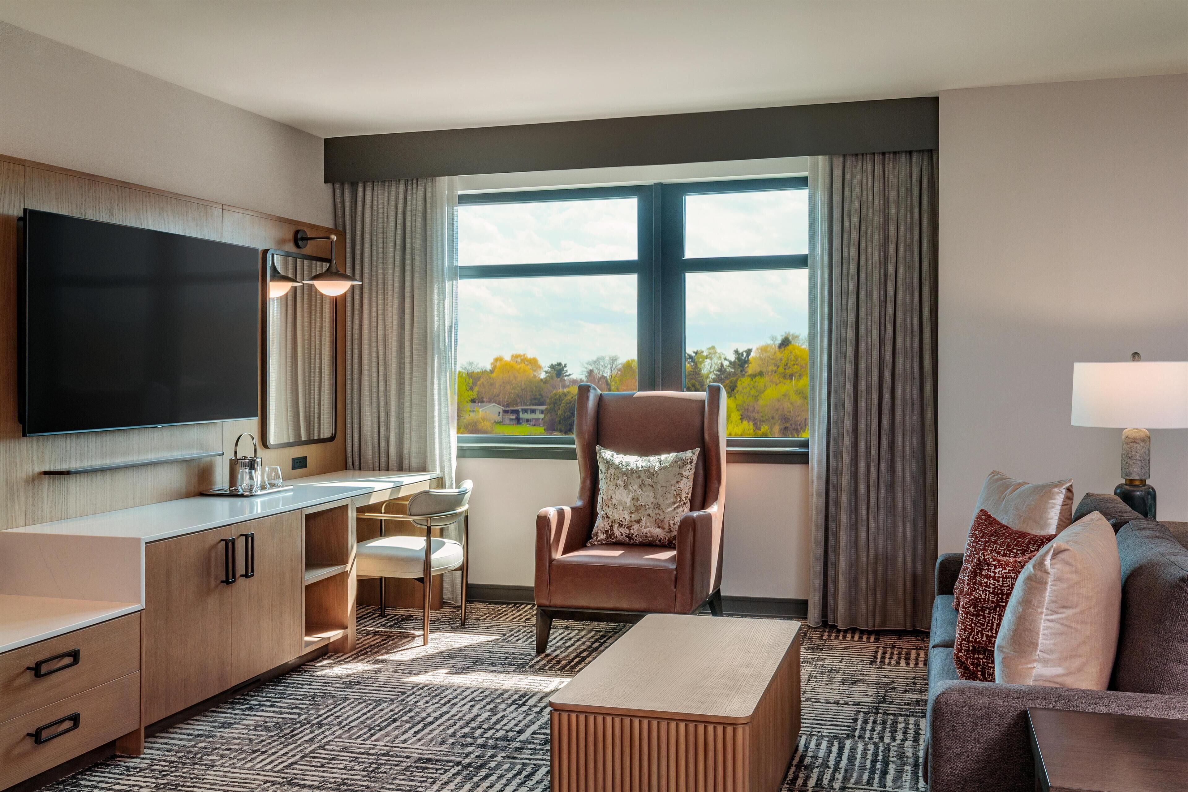 A hotel room with a flat-screen TV, a sofa, a chair, a desk, and a large window with a view.