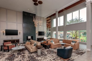 A spacious living area with a large flat-screen TV, multiple seating options, a fireplace, and large windows offering a view of the outdoors. - DoubleTree by Hilton Burlington Vermont (South Burlington)