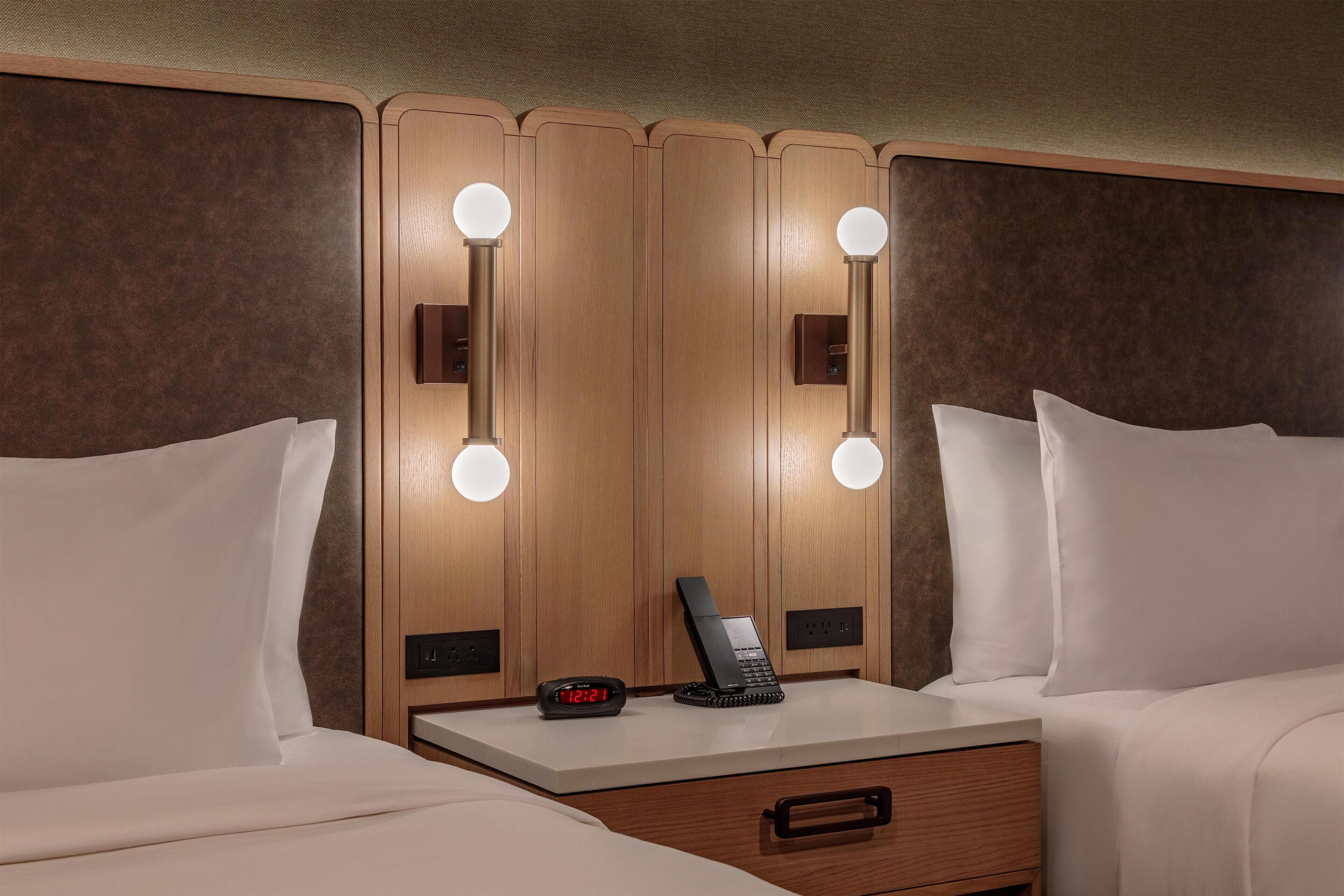 A hotel room with a wooden headboard, bedside tables, a nightstand, a drawer, and a mirror with lights.