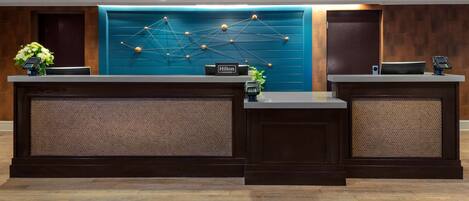 A hotel reception desk with a modern design, featuring a blue wall with a geometric light fixture, two reception counters, and a sign that reads "Hilton Honors".