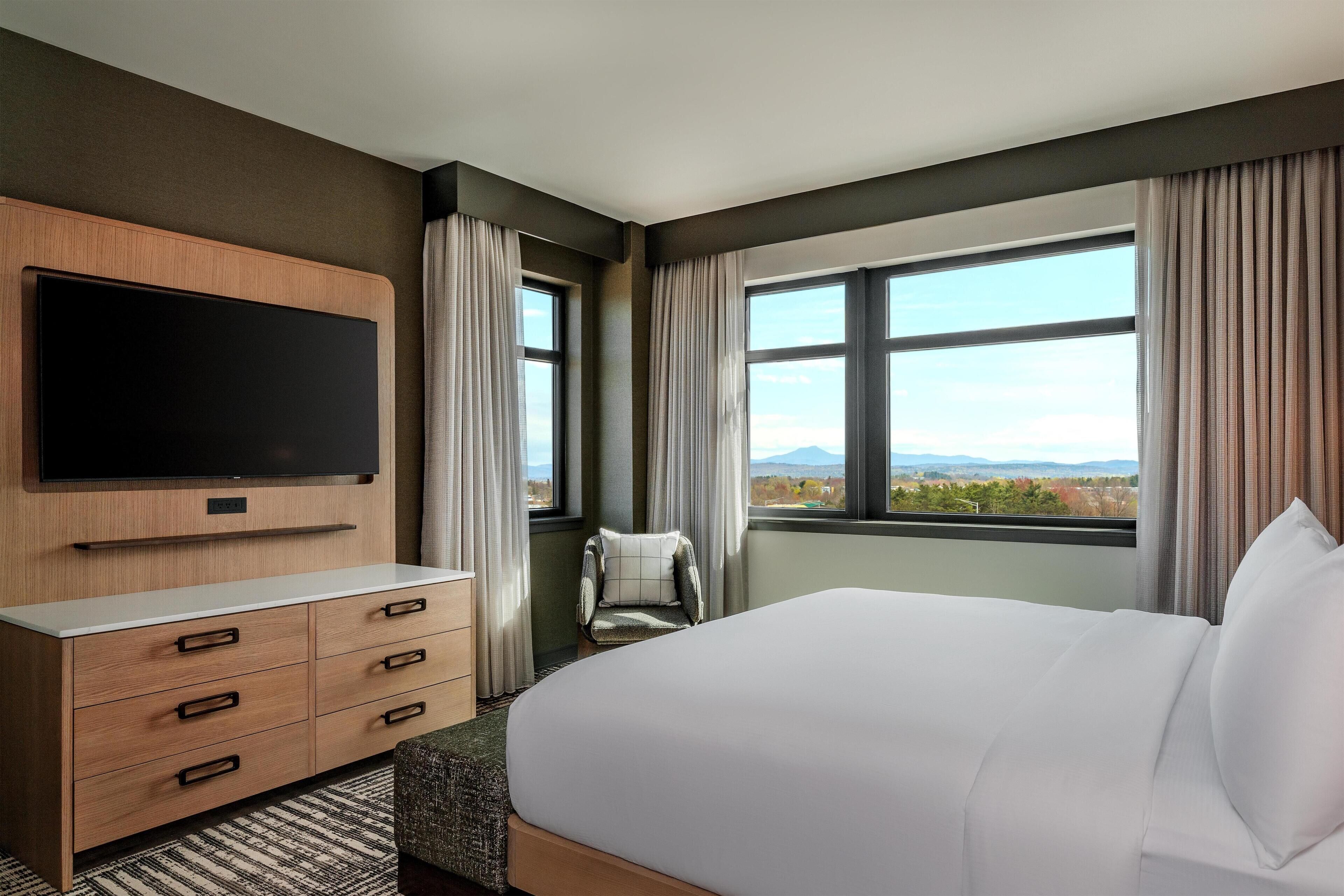 A hotel room with a bed, a TV mounted on the wall, a chair, and a large window offering a view.