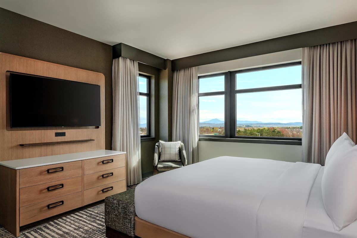 Une chambre d’hôtel avec un lit, un téléviseur fixé au mur, une chaise et une grande fenêtre donnant sur un paysage.