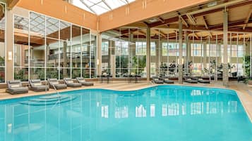 A large indoor swimming pool with lounge chairs and tables, surrounded by large windows and wooden beams.