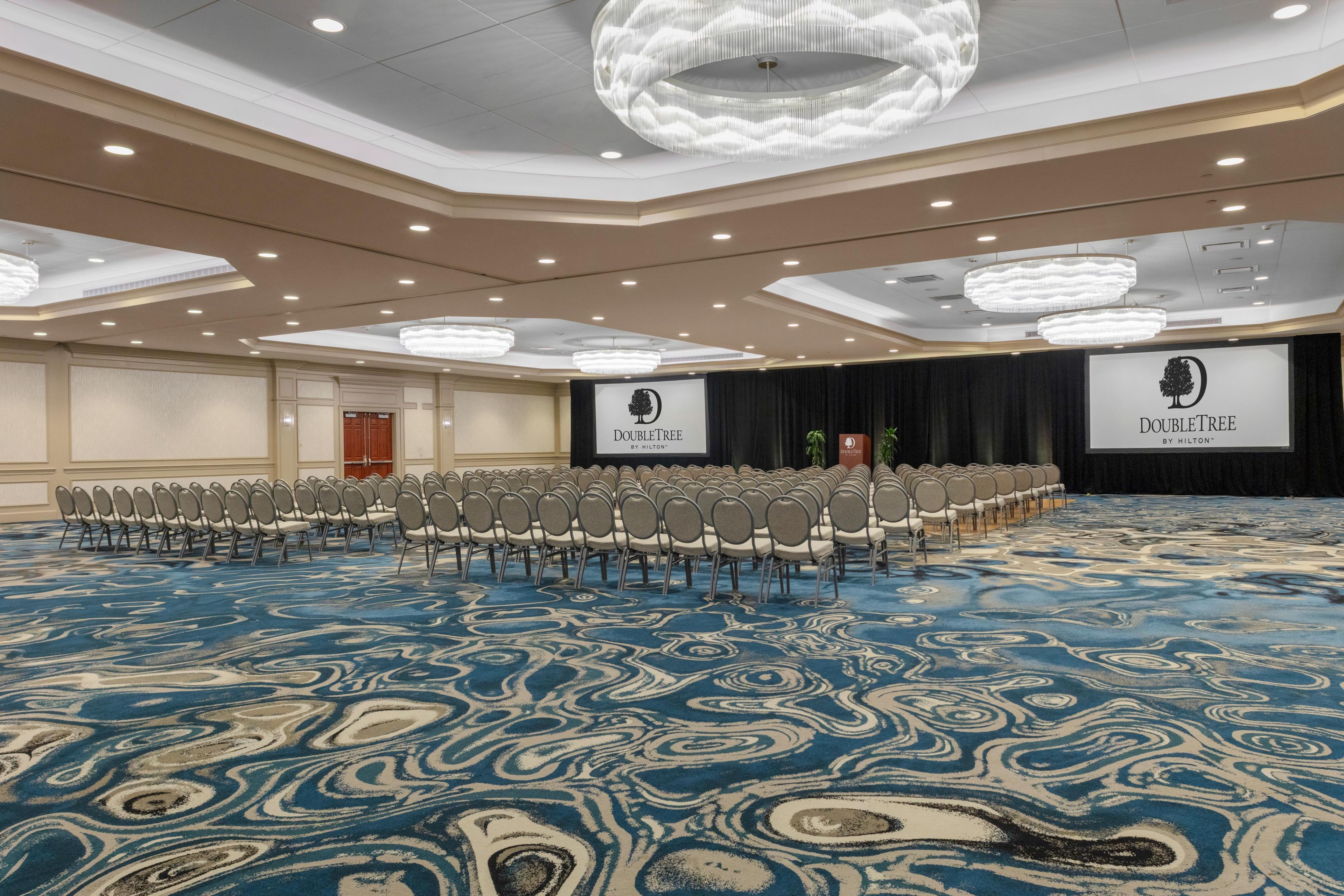 Un salón de conferencias con filas de sillas, un escenario con un atril y una pantalla grande mostrando el logotipo de DoubleTree.