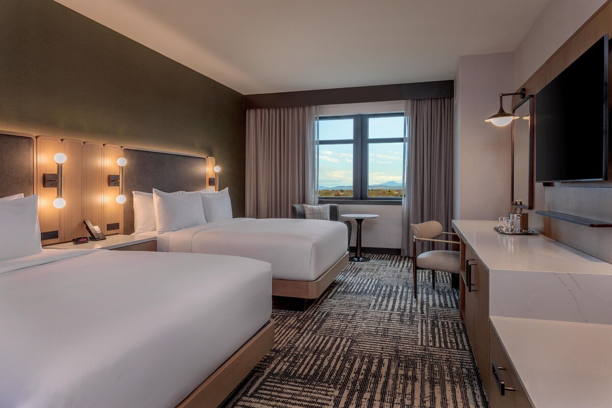 A hotel room with two beds, a desk, a chair, a TV, and a window with a view.