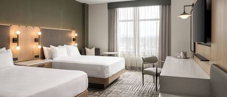 A hotel room with two beds, a desk, a chair, a TV, and a large window with curtains.