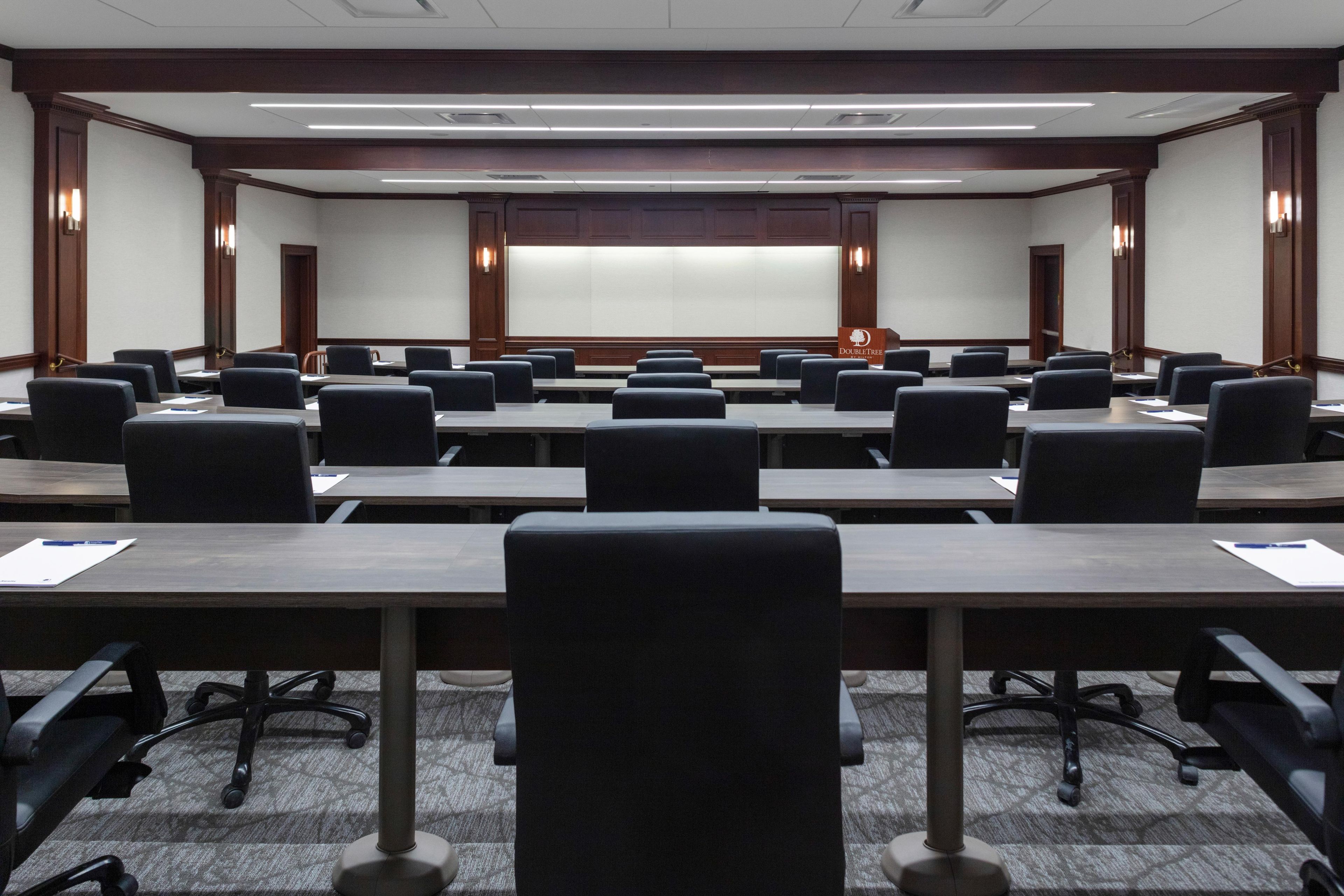 A conference room with multiple tables and chairs, a podium, and a large screen at the front.