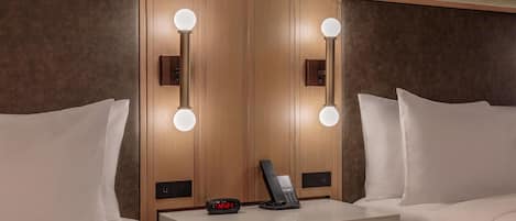 A hotel room with a wooden headboard, bedside tables, a nightstand, a drawer, and a mirror with lights.