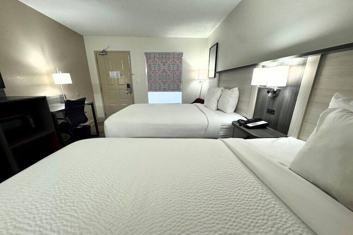 A hotel room with two beds, a desk with a chair, a TV, a lamp, and a door with a patterned curtain.