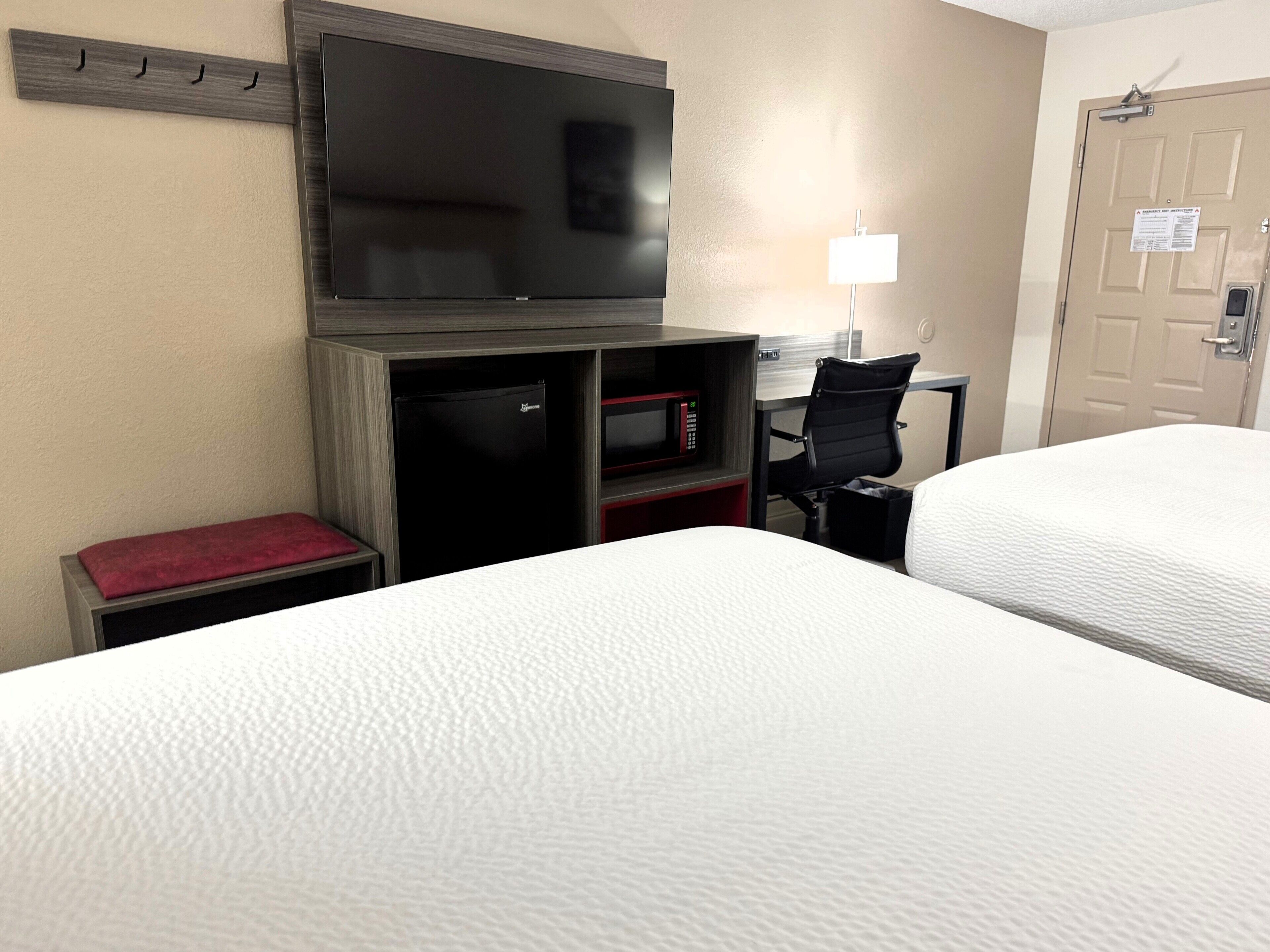 Une chambre d’hôtel avec deux lits, un téléviseur à écran plat fixé au mur, un bureau avec un ordinateur, un four à micro-ondes et une chaise.