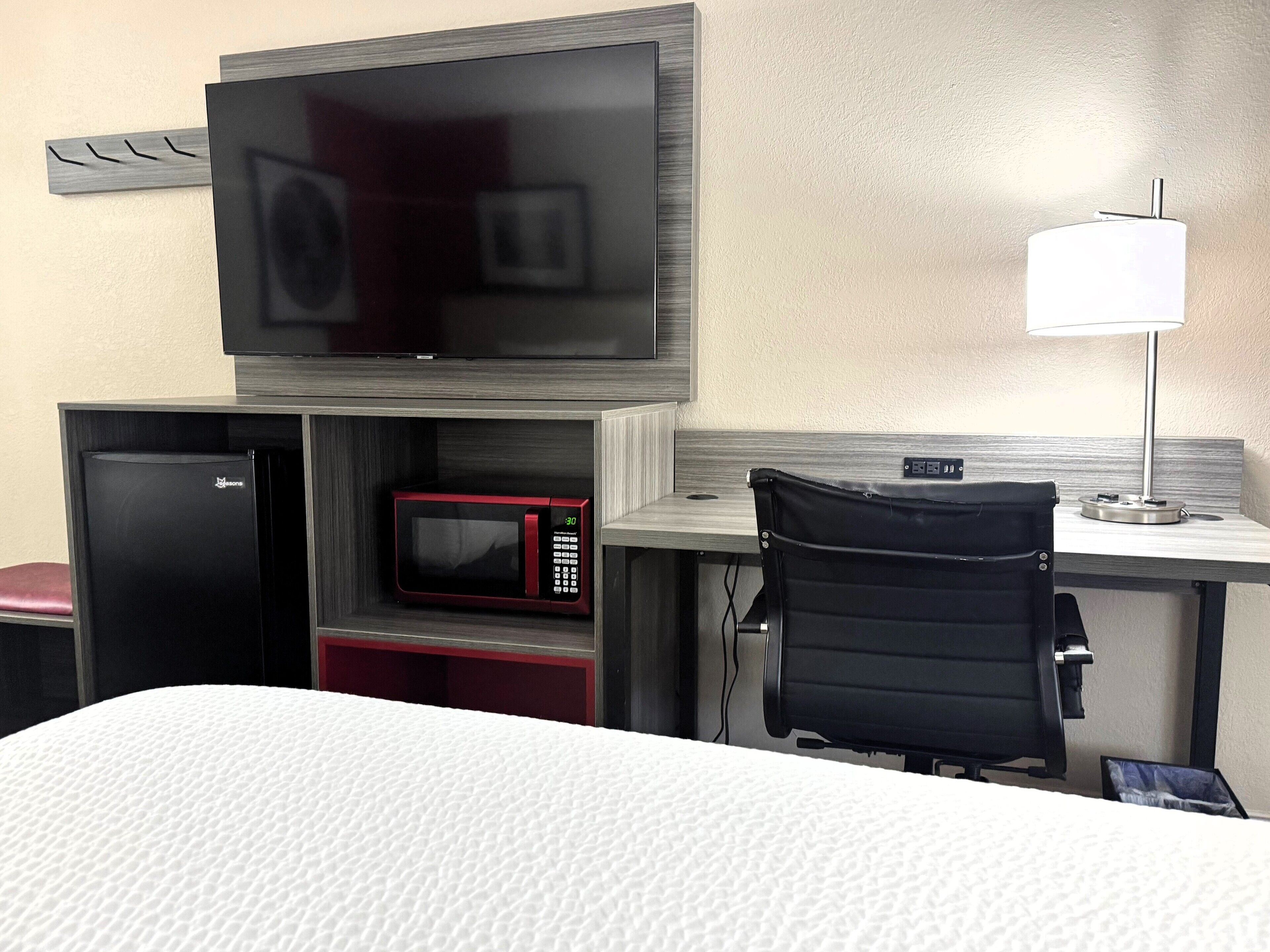 Une chambre d’hôtel équipée d’un téléviseur fixé au-dessus d’un bureau, d’un four à micro-ondes placé en dessous, d’un réfrigérateur sur le côté gauche et d’une chaise devant le bureau.