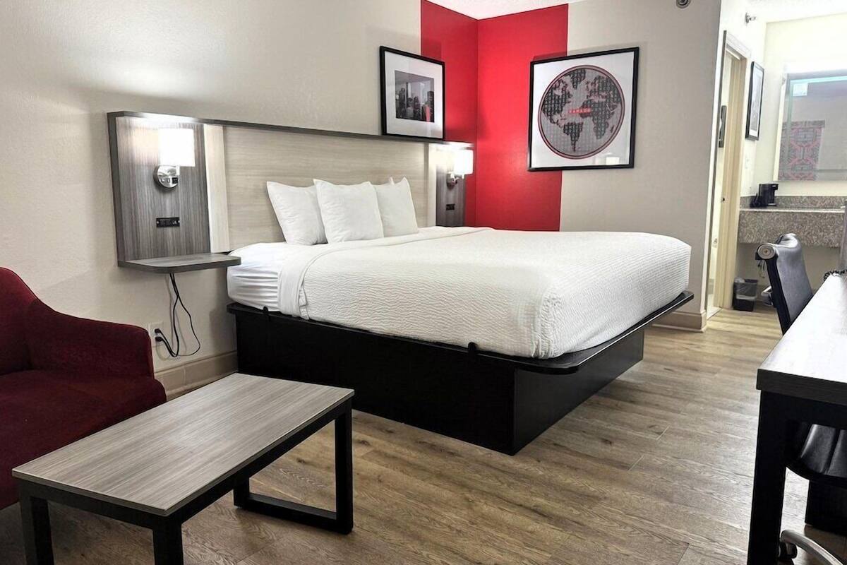 A hotel room with a bed, a desk, a chair, a red accent wall, and framed pictures on the wall.