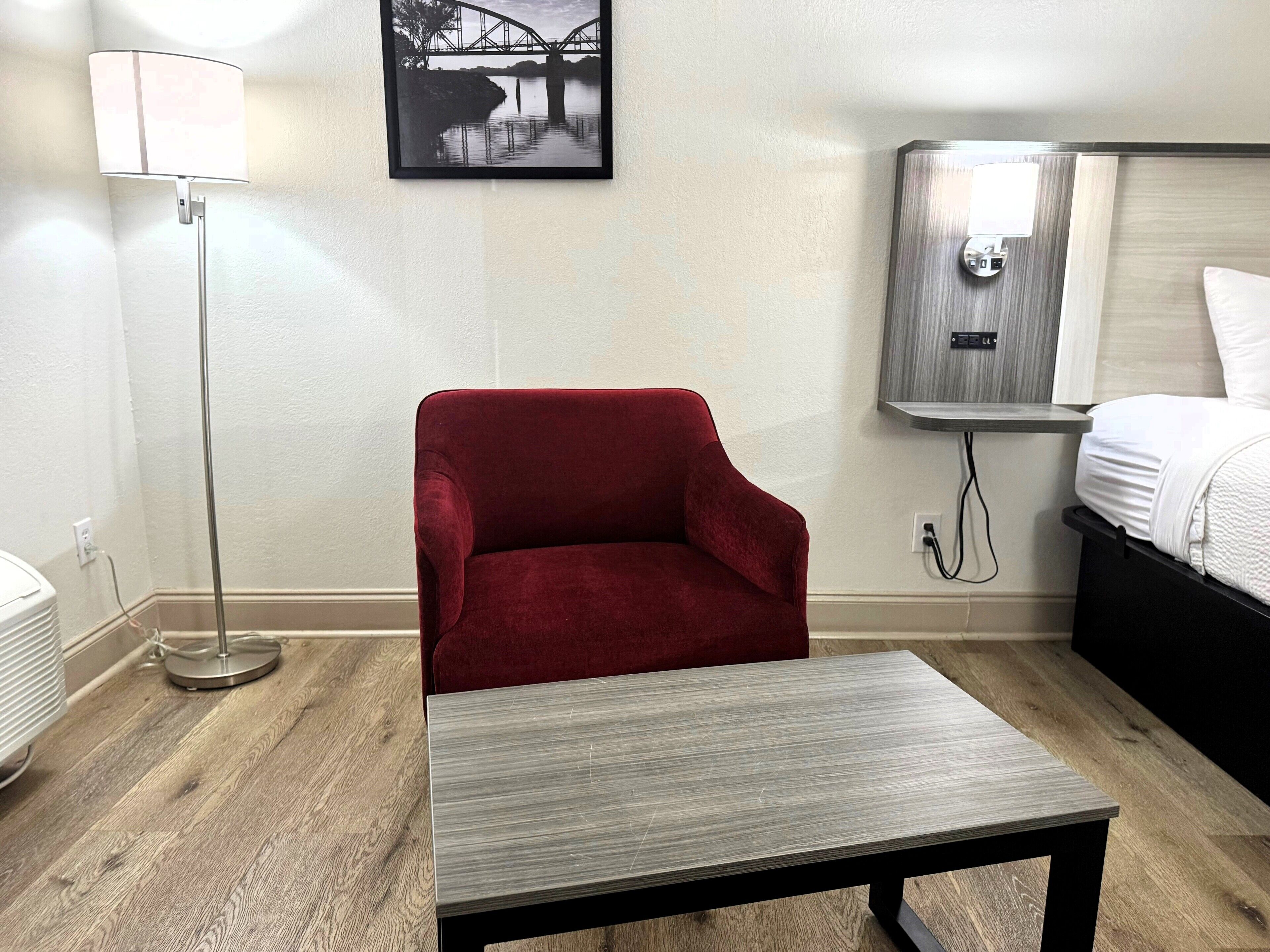 A hotel room with a red armchair, a small table, a bed, a nightstand, a lamp, and a framed picture on the wall.