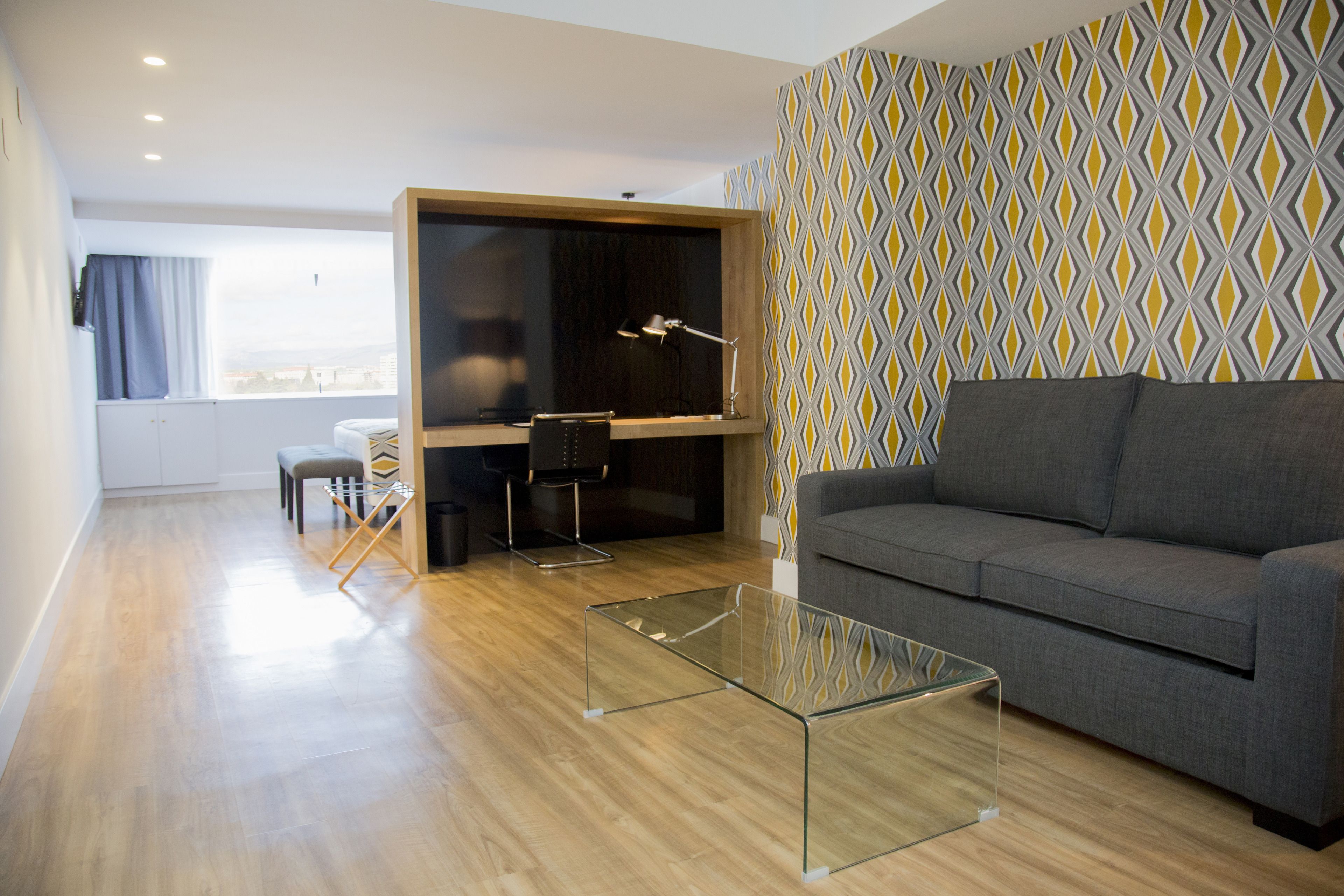 A modern living room with a grey sofa, a glass coffee table, and a wooden floor. There is a desk with a chair and a lamp. The room has a window with curtains and a patterned wallpaper.