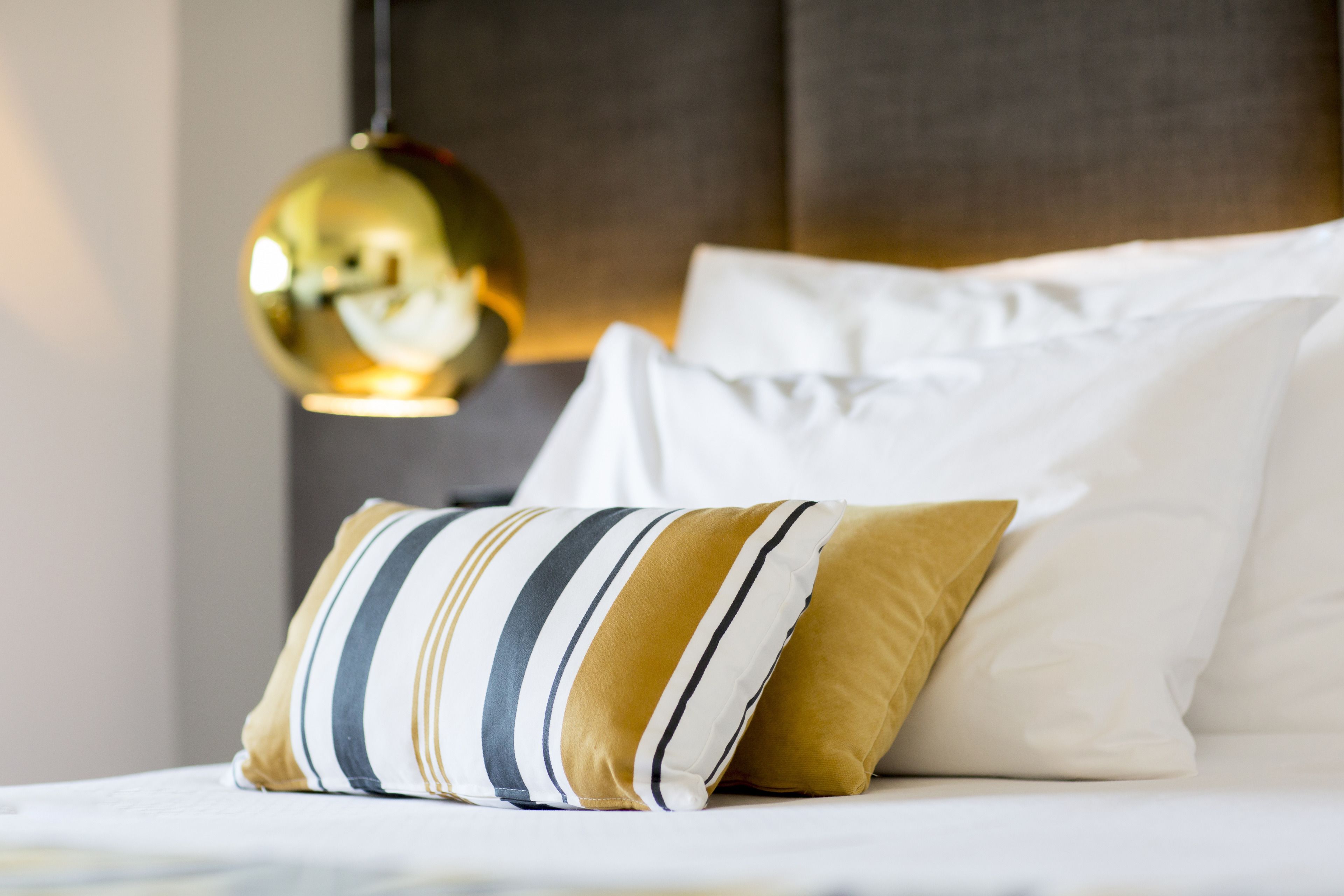 A neatly made bed with white and striped pillows, a gold pendant light, and a dark headboard.