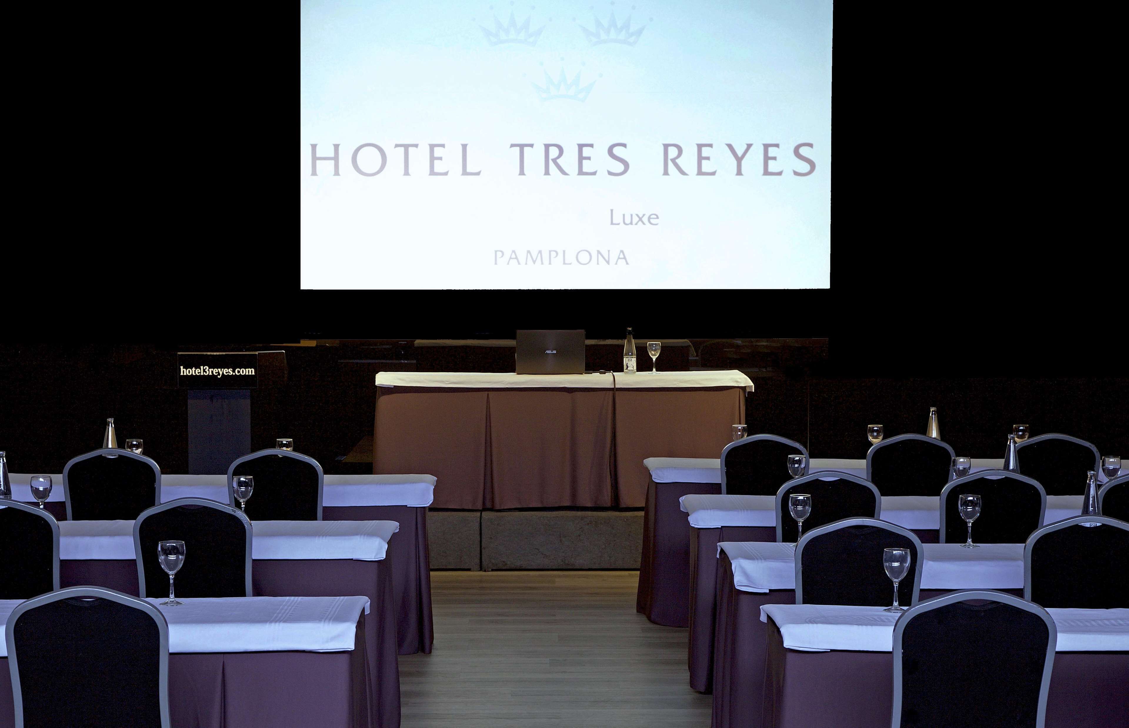 Una sala de conferencias en Hotel Tres Reyes, Luxe Pamplona, con filas de mesas y sillas, un atril y una pantalla de proyección mostrando el nombre del hotel.