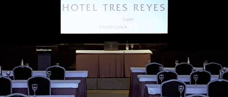 A conference room at Hotel Tres Reyes, Luxe Pamplona, with rows of tables and chairs, a podium, and a projection screen displaying the hotel's name.