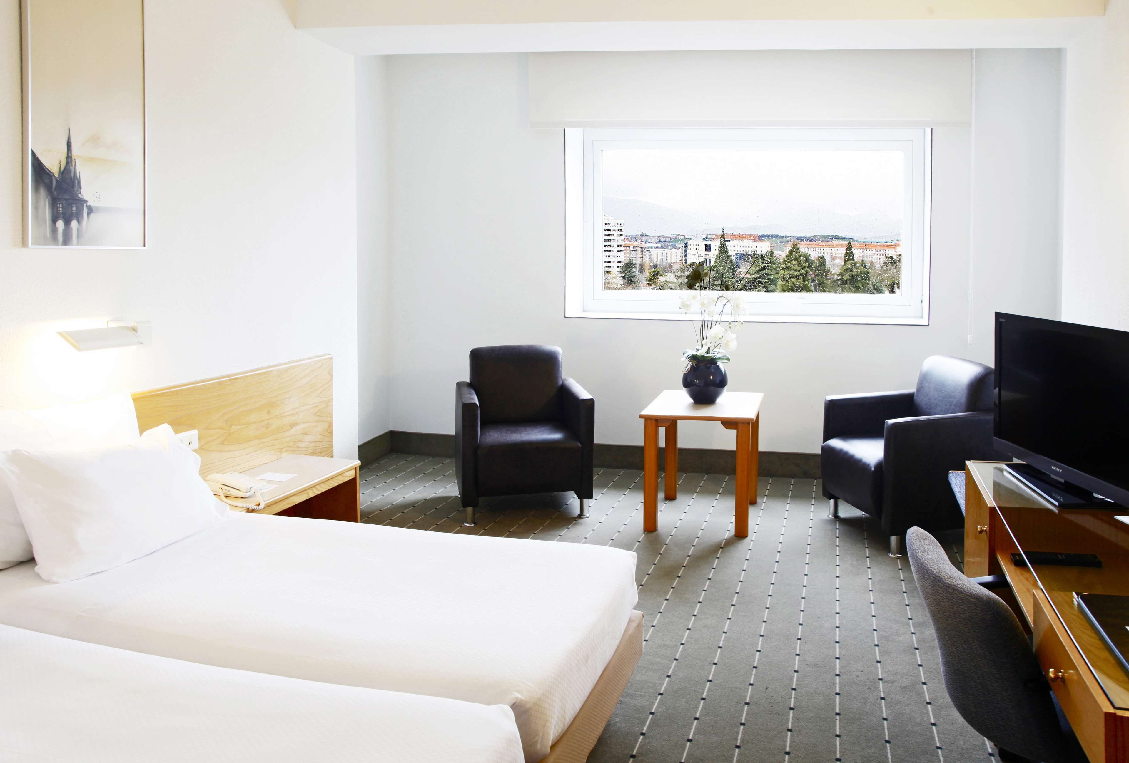 Habitación de hotel con cama, dos sillones, una mesita con un florero, televisión y ventana con vista a la ciudad.