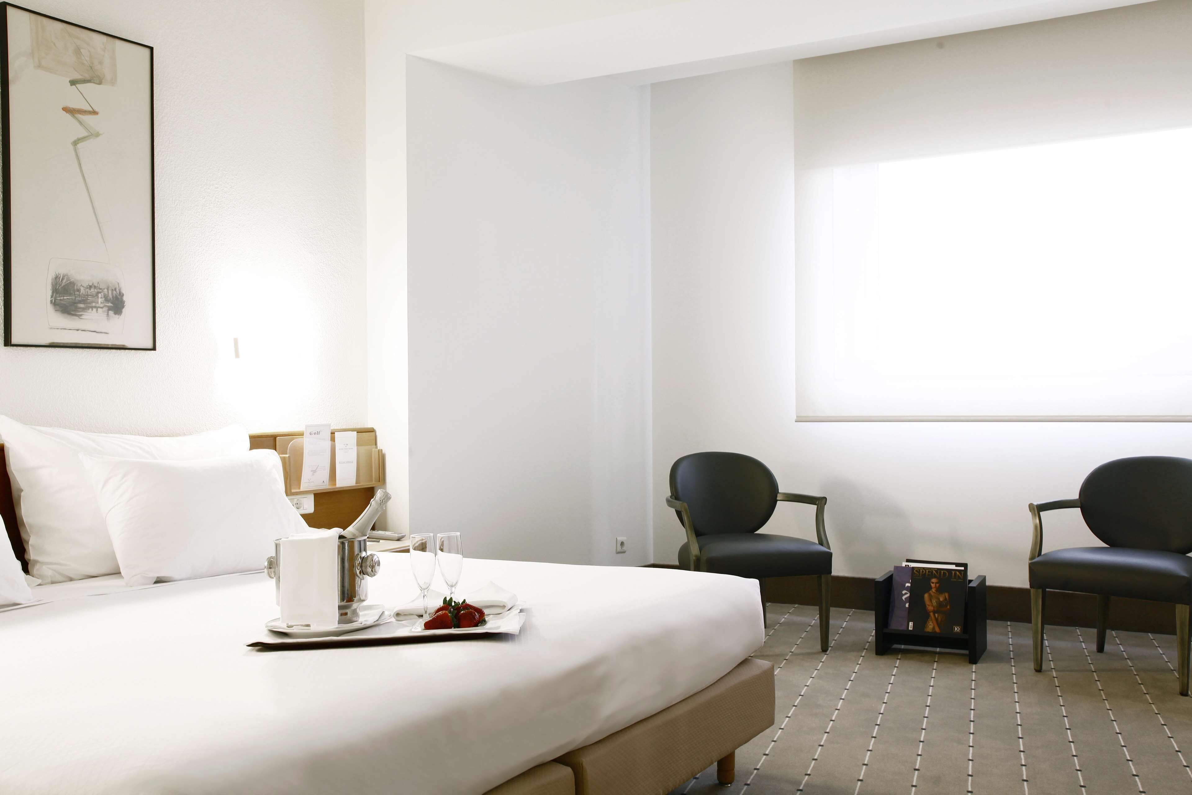A hotel room with a bed, two chairs, a tray with food and drinks, and a bookshelf.