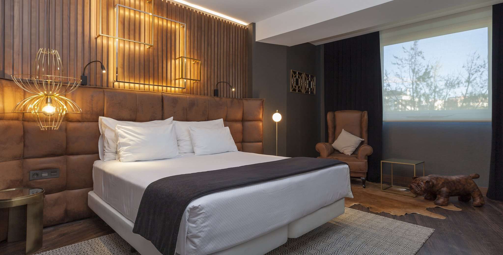 Un dormitorio moderno con una cama grande, cabecera de madera, un sillón de cuero, una mesita y un perro.