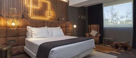 A modern bedroom with a large bed, a wooden headboard, a leather armchair, a small table, and a dog.