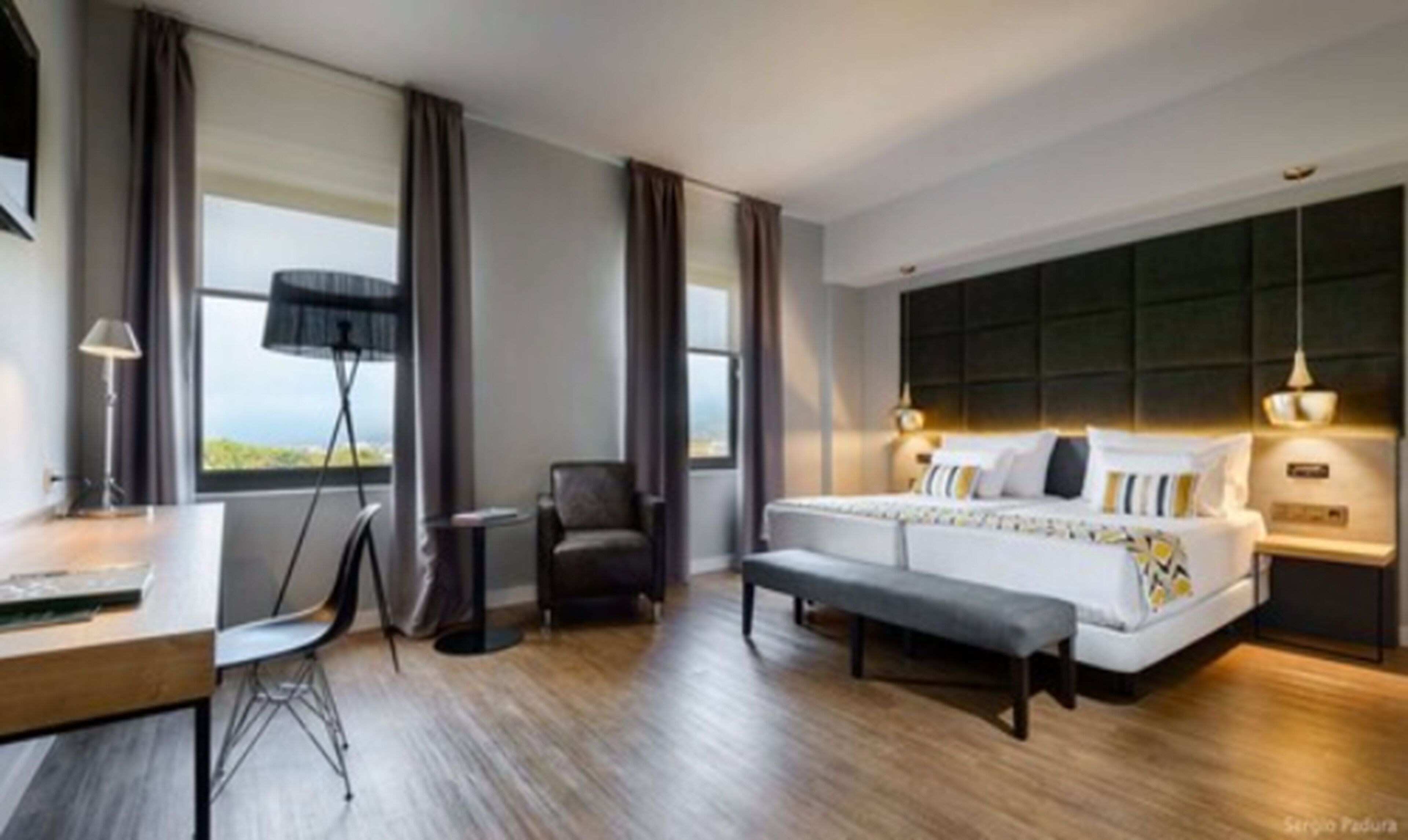 A modern hotel room with a large bed, a desk, and a chair. There are windows with curtains, a view of the outdoors, and a headboard with built-in lighting.