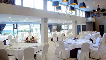Un amplio salón de banquetes con varias mesas redondas dispuestas para comer, sillas blancas y un techo alto con lámparas modernas.