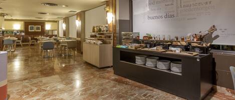 Zona de desayuno de hotel con barra de buffet, área de asientos y una pantalla mostrando saludos en varios idiomas.
