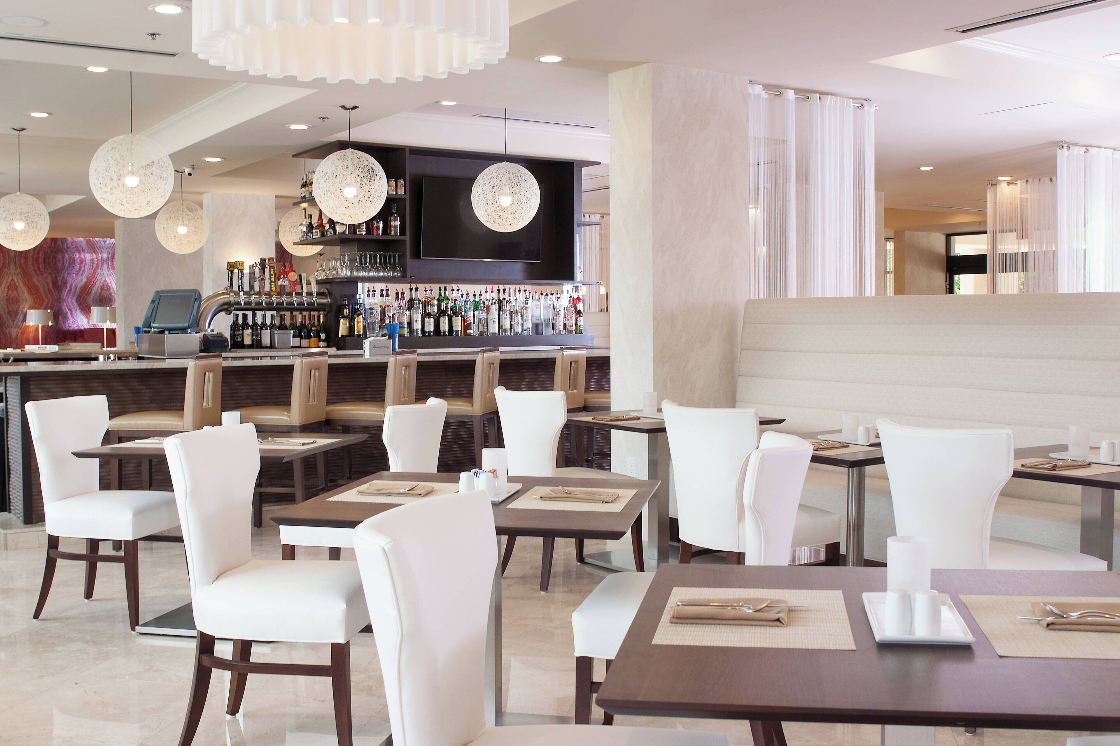 A modern restaurant with white chairs and tables, a bar area with a variety of bottles, and pendant lighting.