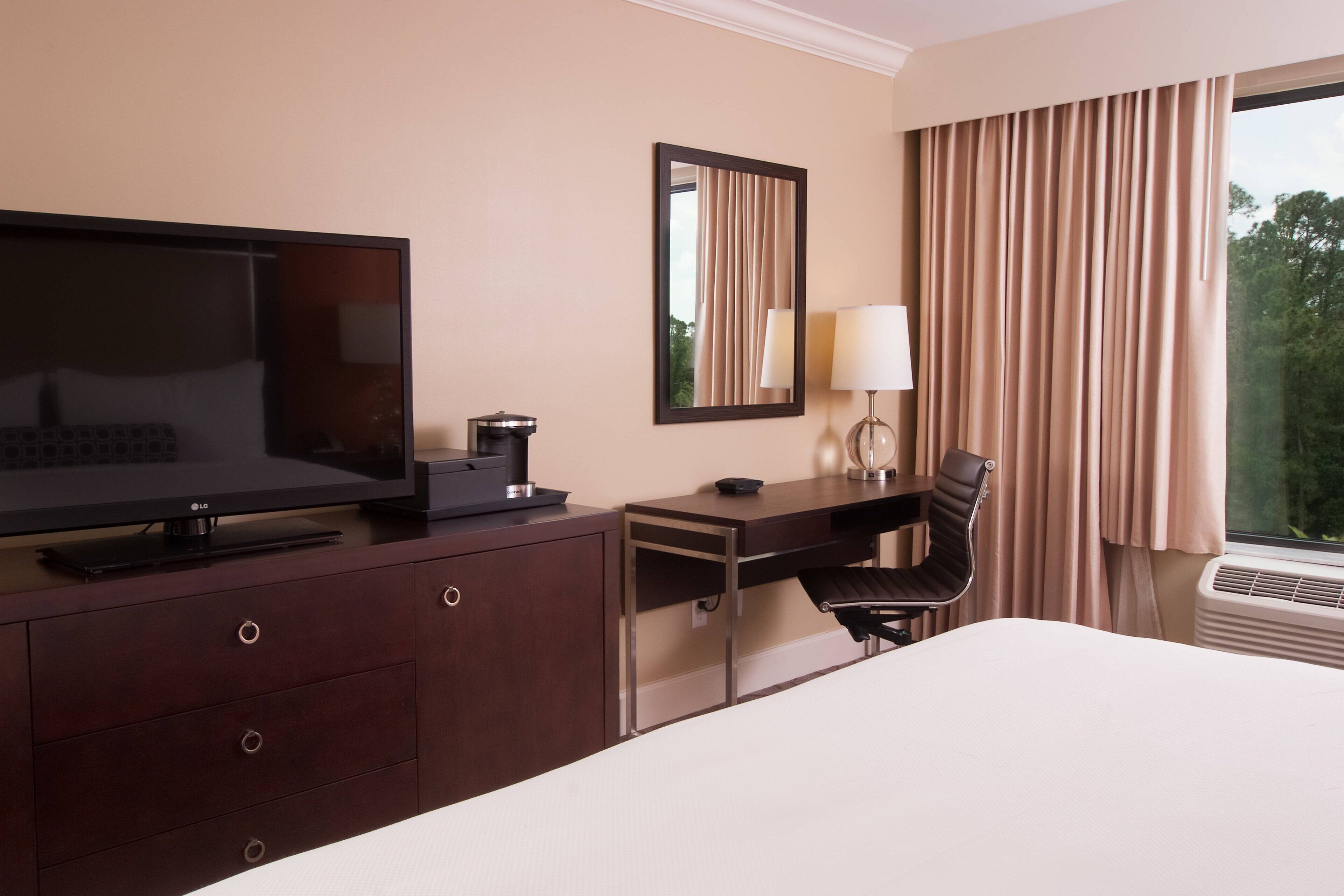 Une chambre d’hôtel comprenant un lit, un bureau avec une chaise, une télévision, un miroir, une lampe et une fenêtre avec des rideaux.