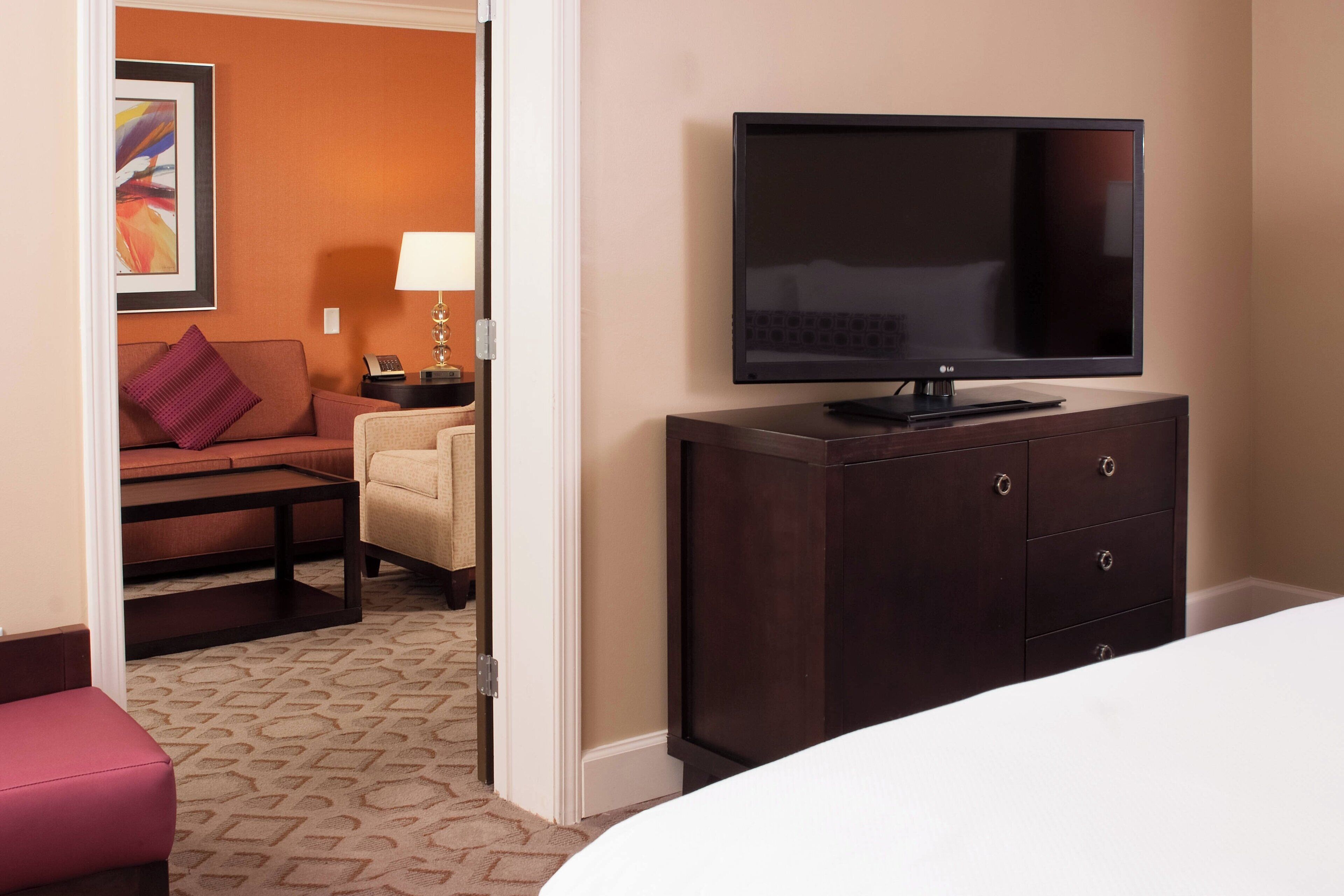 A hotel room with a dark wooden dresser, a flat-screen TV, a beige sofa, and a red armchair.