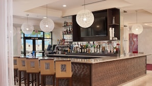 A bar area with a counter, multiple bar stools, a television mounted above the bar, and a variety of bottles and glasses on the shelves.