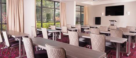 A conference room with multiple tables and chairs, large windows with curtains, a flat-screen TV, and a projector.