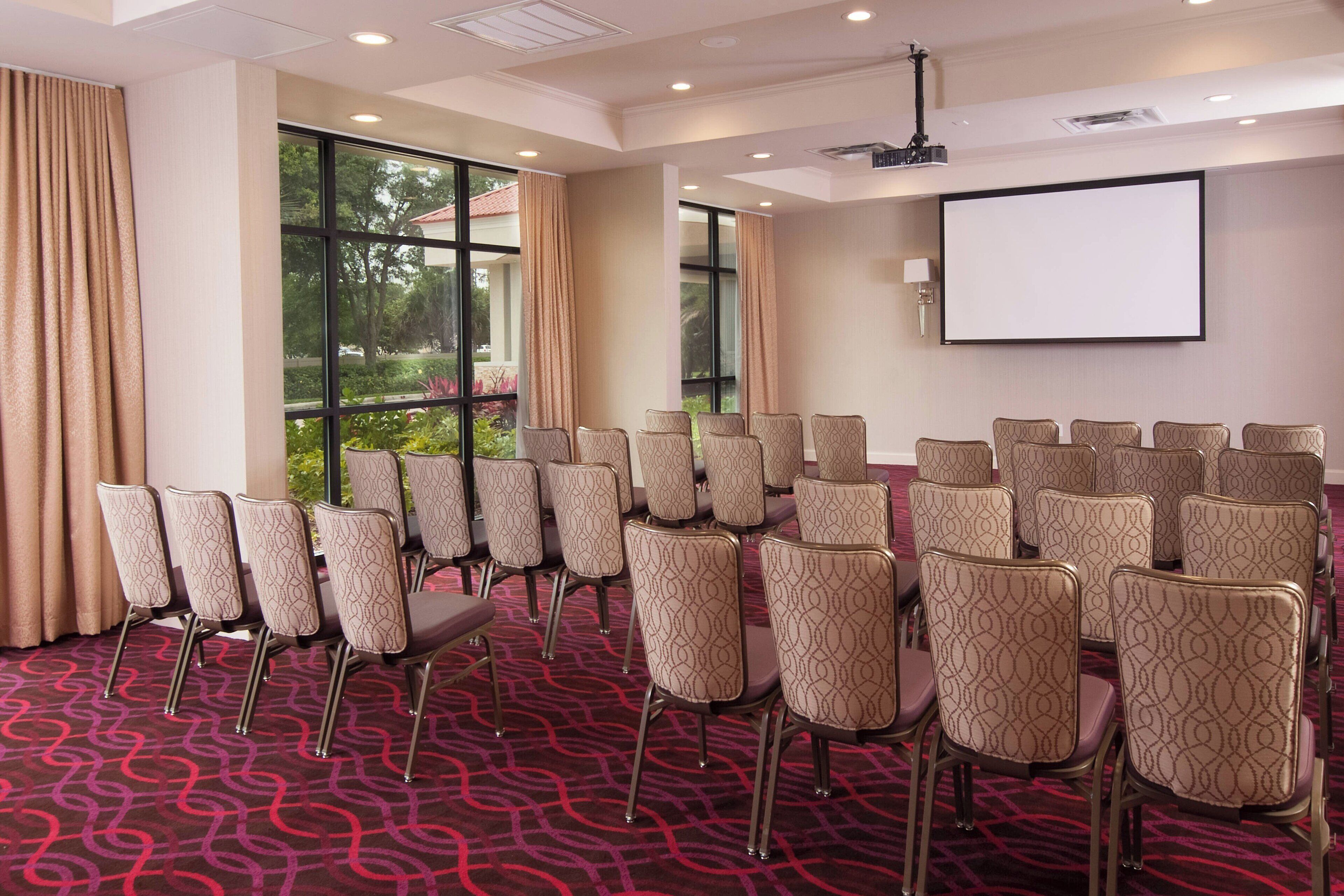 A conference room with rows of chairs, a projector screen, and large windows with curtains.
