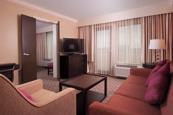A hotel room with a sofa, a small table, a TV, a lamp, and a window with curtains.