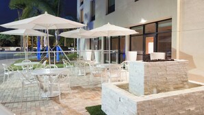 An outdoor patio with white tables and chairs, umbrellas, and a stone planter with a water feature.