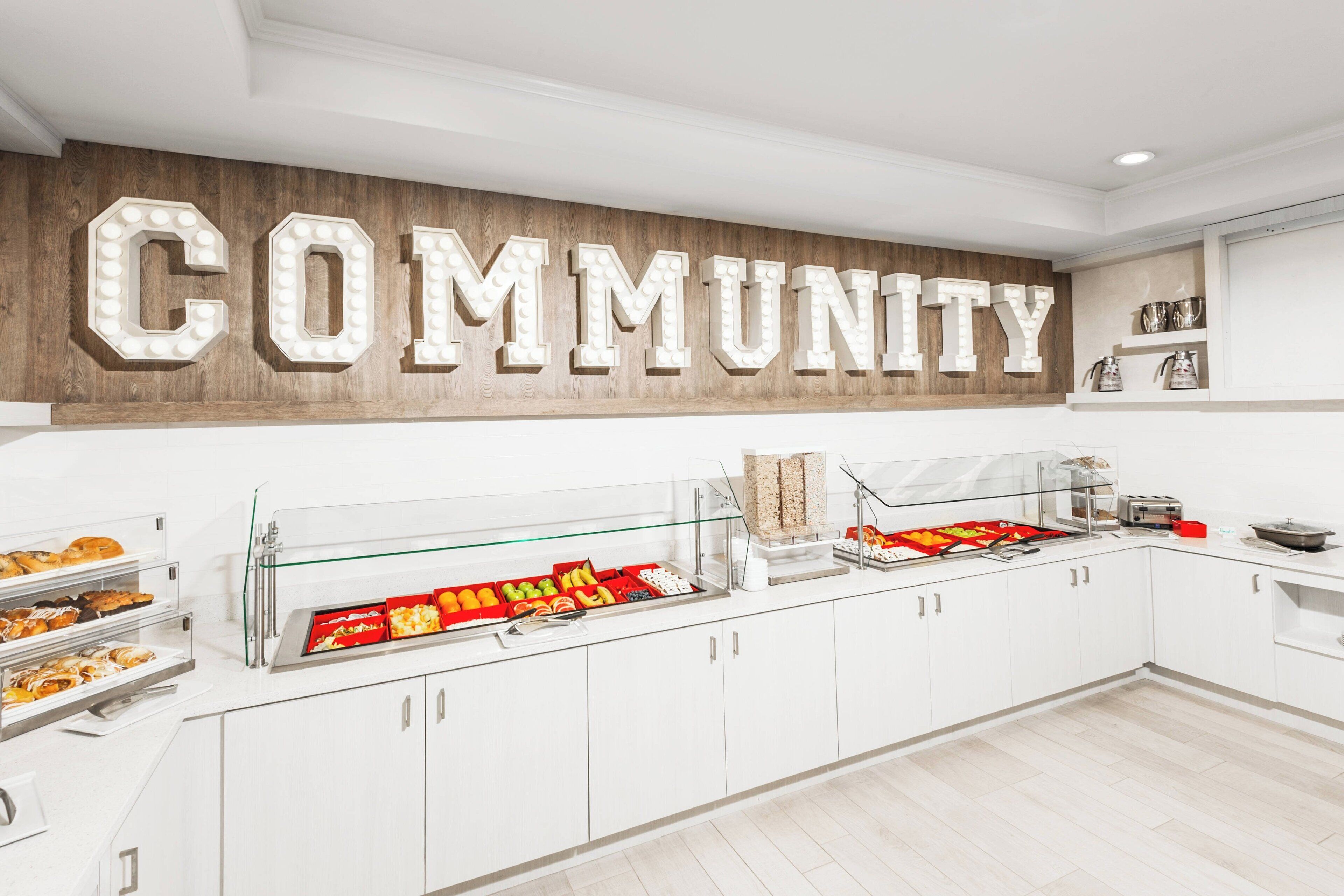 Cozinha moderna com parede de madeira exibindo a palavra "COMMUNITY" em letras iluminadas, um buffet com diversos alimentos e armários brancos.