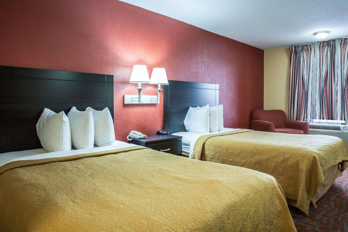 Quarto de hotel com duas camas, uma mesa de cabeceira com telefone, uma cadeira e uma janela com cortinas.