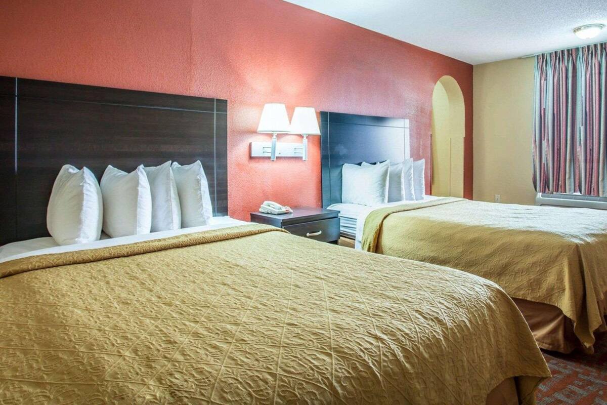 Quarto de hotel com duas camas, uma mesa de cabeceira, uma luminária e uma janela com cortinas.
