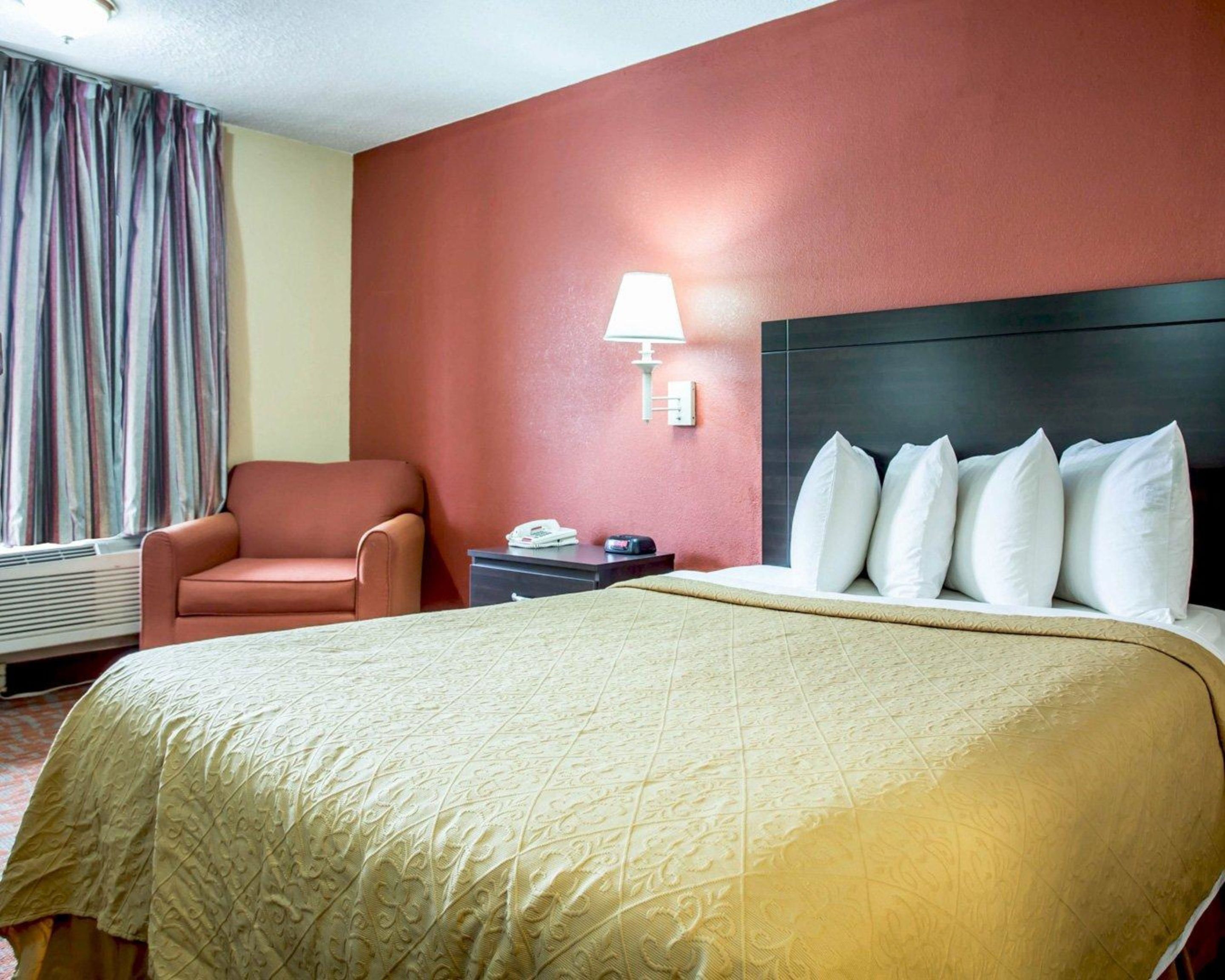 Quarto de hotel com uma cama grande, uma parede de destaque vermelha, uma cadeira, uma mesa de cabeceira com abajur e uma janela com cortinas.