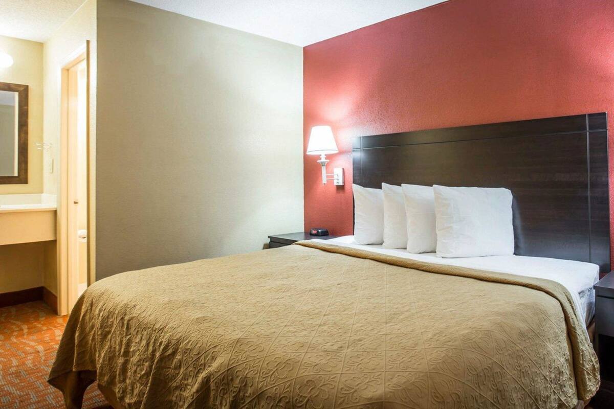 Quarto de hotel com uma cama grande, uma mesinha de cabeceira, um abajur e uma parede em tom de vermelho.