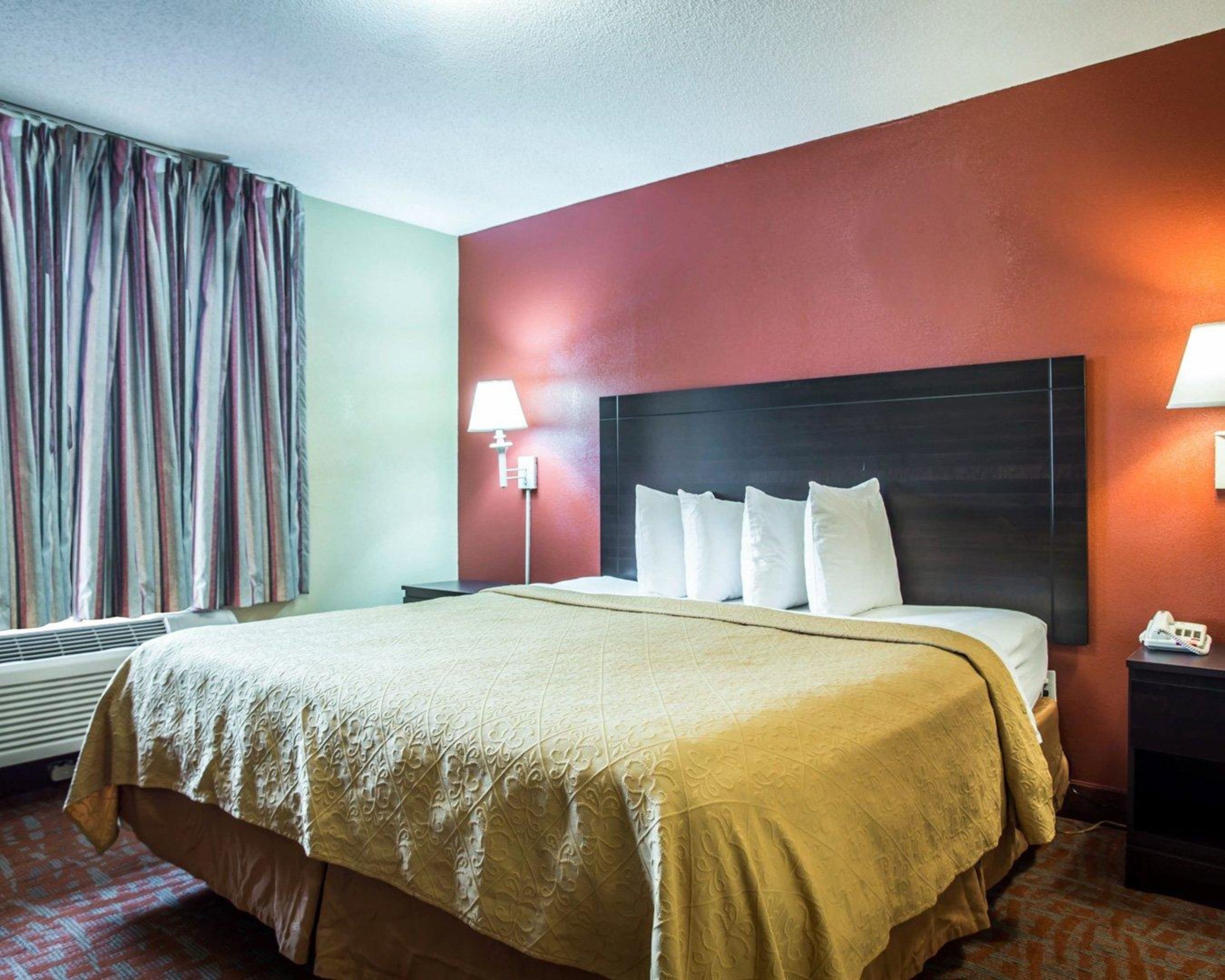 A hotel room with a large bed, a nightstand with a lamp, a telephone, and a window with curtains.