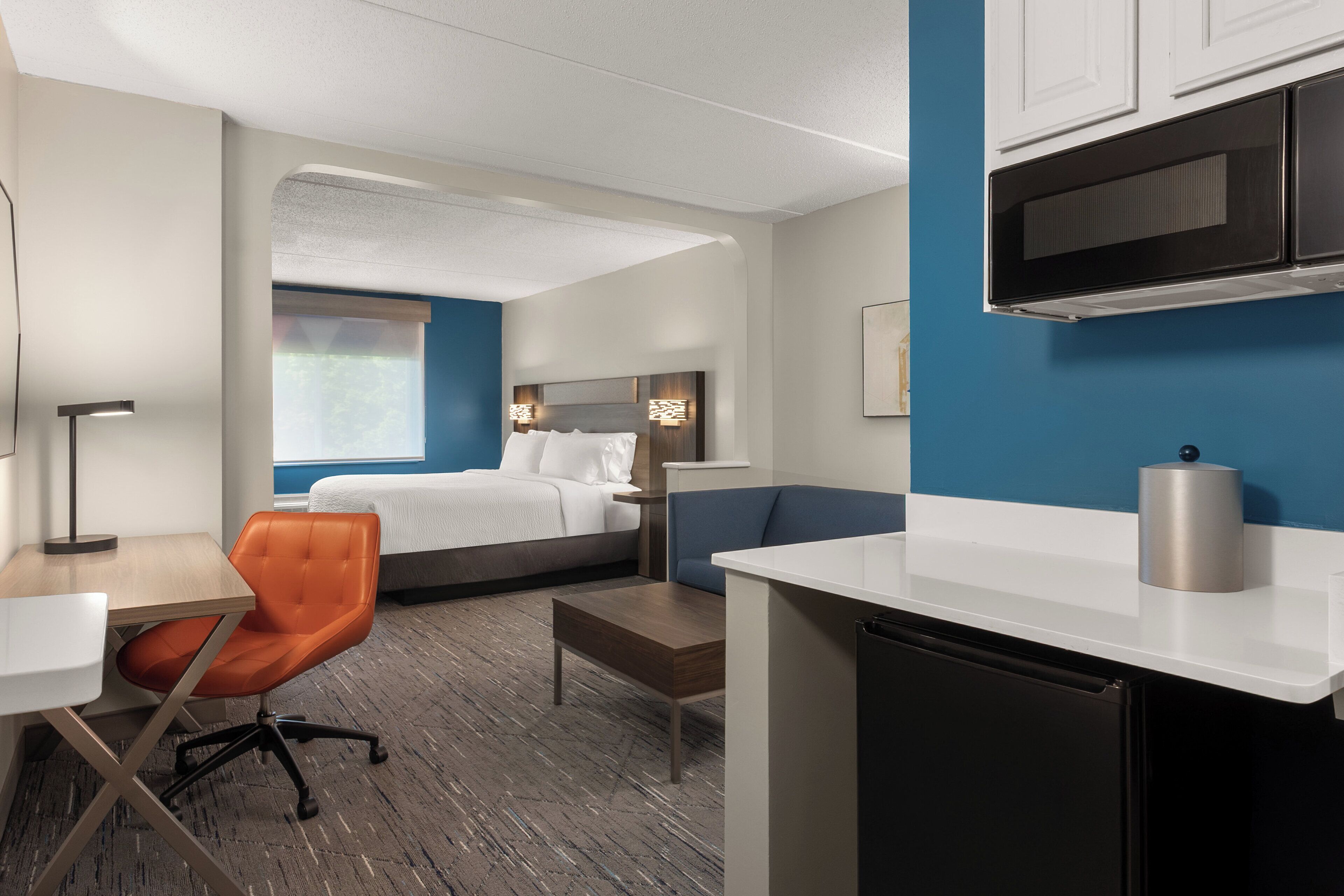 A hotel room with a bed, a desk, a chair, a kitchenette, and a blue accent wall.