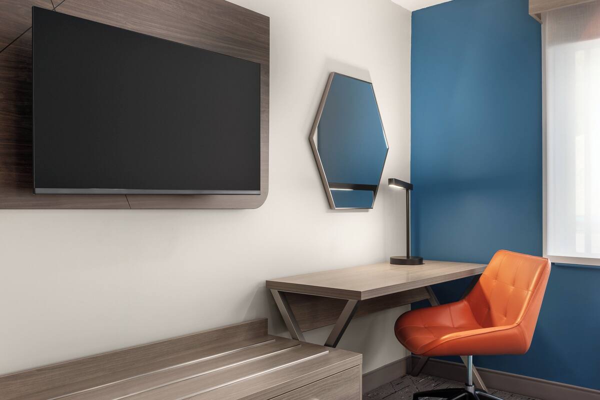 A modern room with a large flat-screen TV mounted on the wall, a desk with a chair, and a window with blinds.