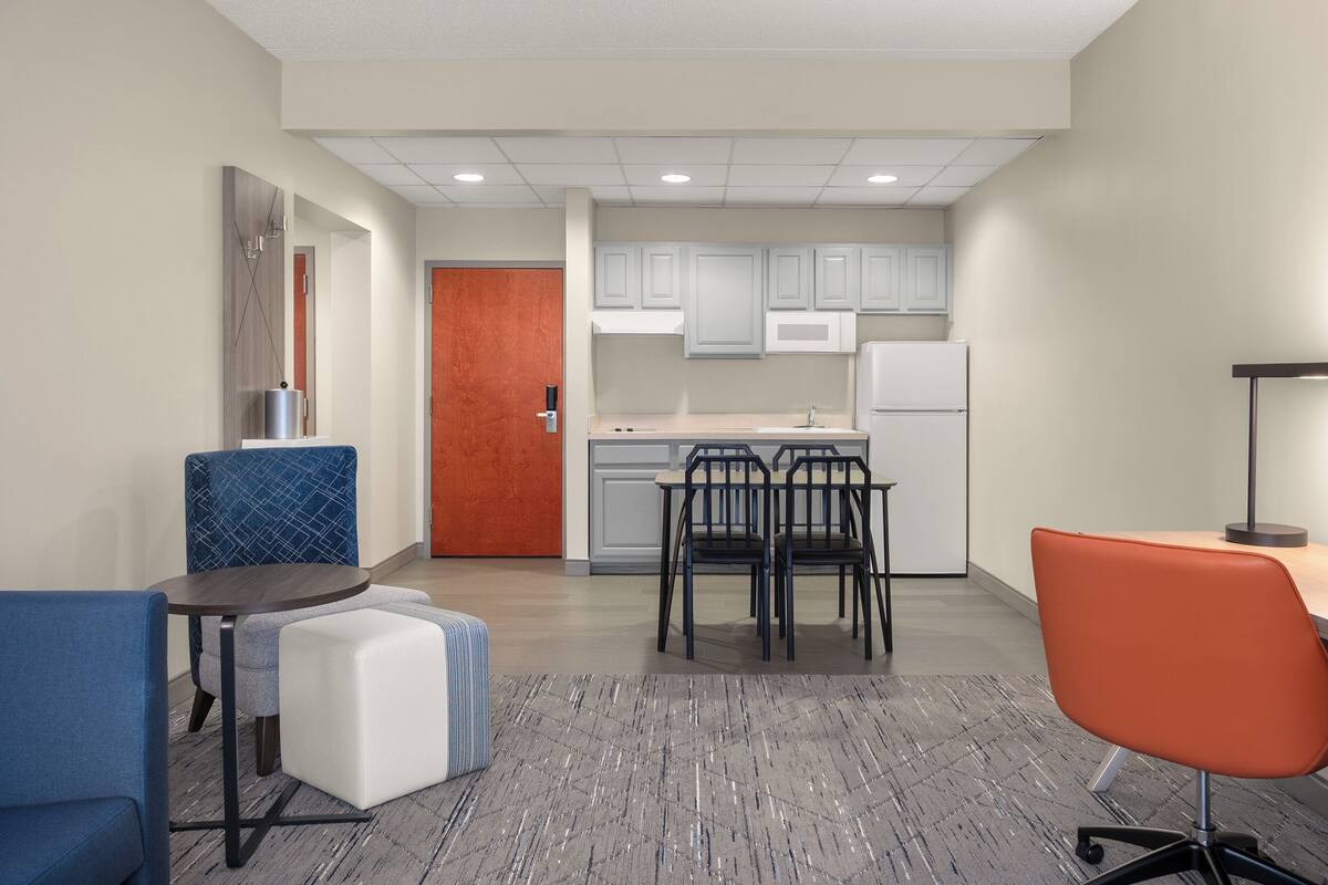 A modern kitchen with white cabinets, a refrigerator, and a microwave. There are two dining tables with chairs, one with a blue chair and the other with an orange chair. A small white ottoman is placed between the tables.